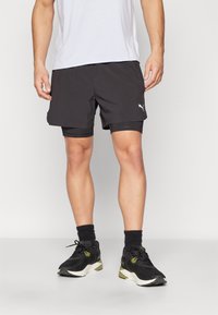 Pantalones cortos deportivos negros con bolsillos laterales y un logo de Puma en blanco, combinados con zapatillas negras con detalles olivo y calcetines oscuros.