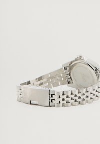 Ceas din oțel inoxidabil argintiu cu un design de brățară legată. Dispune de un carcasă rotundă, o suprafață texturată și un sistem de închidere tip cataramă.