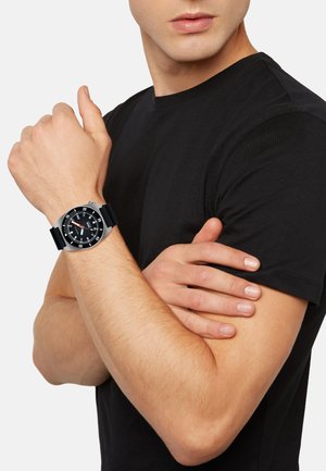Montre noire avec un cadran rond, accents argentés et un bracelet en caoutchouc noir, portée au poignet, à côté d'un t-shirt noir.