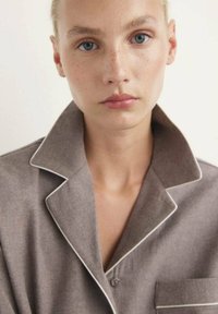 Joven mujer con ojos azules y pecas lleva una camisa gris con cuello y ribetes blancos, mirando directamente hacia adelante contra un fondo plano.