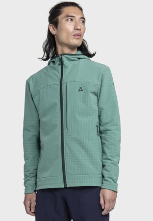 Mann mit langen dunklen Haaren, der einen strukturierten grünen Kapuzenpullover mit Reißverschluss trägt und zur Seite schaut vor einem schlichten hellen Hintergrund.