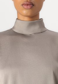Top grigio chiaro a collo alto realizzato in un tessuto morbido simile alla seta, con una silhouette aderente e una leggera lucentezza, caratterizzato da cuciture minime.