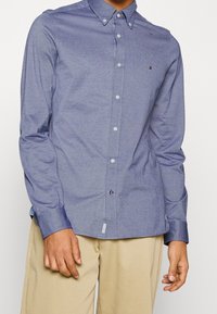 Camicia con bottoni a maniche lunghe in tessuto chambray blu scuro, con colletto, bottoni bianchi e un piccolo logo sulla tasca del petto.