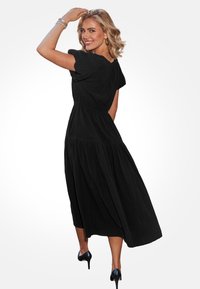 Czarna maxi sukienka z lekkiego materiału, z krótkimi bufkami, dopasowaną talią i spódnicą z falbanami, stylizowana z wysokimi obcasami.