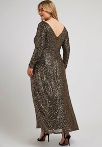 Lång, glittrande klänning med långa ärmar, V-back design och skimrande guld- och svarta paljetter. Stylad med svarta högklackade sandaler.