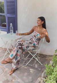 Jumpsuit bez ramiączek, w deseń z motywami tropikalnymi w odcieniach różu, zieleni i beżu, w połączeniu z brązowymi klapkami, siedzący przy jasnym stole.