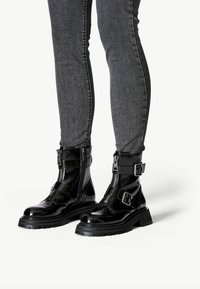 Botas de tobillo de charol negro con hebillas ajustables, cremalleras laterales y una suela de goma gruesa; combinadas con jeans ajustados de mezclilla oscura.