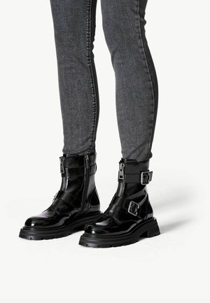 Botas de tobillo de charol negro con hebillas ajustables, cremalleras laterales y una suela de goma gruesa; combinadas con jeans ajustados de mezclilla oscura.