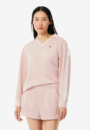 Joven mujer con cabello largo y oscuro, vestida con una camiseta de manga larga rosa claro con rayas blancas y pantalones cortos a juego sobre un fondo liso.