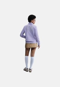 Felpa zip-up lilla con il logo "Nike" sulla schiena, abbinata a una gonna beige, calze bianche fino al ginocchio e scarpe marroni. Texture liscia.