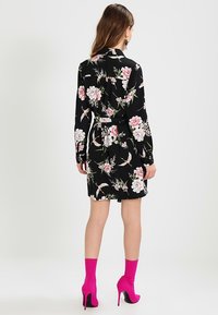 Kvinna klädd i en svart blommig klänning med långa ärmar och ett midjebälte, tillsammans med ljusrosa högklackade ankelskofötter, stående med ryggen vänd mot kameran.