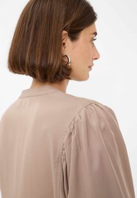 Blusa marrone chiaro con maniche a palloncino, colletto sartoriale e una texture di tessuto liscia. Design semplice che enfatizza colori tenui e dettagli sottili.