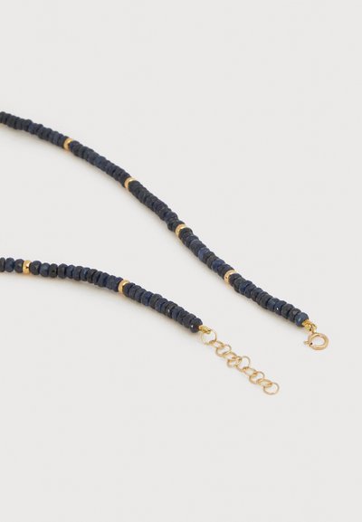 Κολιέ με μικρές μαύρες χάντρες και ομοιόμορφα κατανεμημένες χρυσές λεπτομέρειες, με χρυσή επέκταση αλυσίδας και κούμπωμα με ελατήριο σε λευκό φόντο.