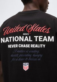 Άτομο που φοράει μαύρο μπλουζάκι με κόκκινο, λευκό και μπλε κείμενο: «Η Εθνική Ανδρών των Ηνωμένων Πολιτειών ποτέ δεν κυνηγά την πραγματικότητα», μαζί με το λογότυπο της ασπίδας των ΗΠΑ.