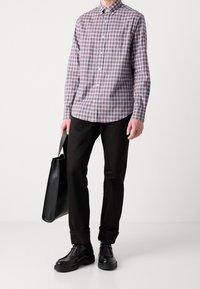 Ein Mann trägt ein rot-weiß kariertes Hemd, schwarze Hosen und schwarze Schuhe. Er hält eine schwarze Leder-Tote-Bag vor einem weißen Hintergrund.