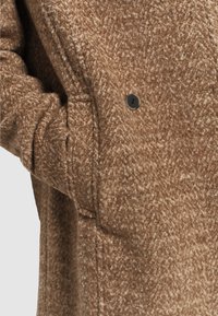 Abrigo de mezcla de lana marrón con una tela texturizada, costuras visibles, bolsillos laterales y un detalle de botón negro redondo.