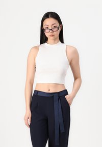 Bijela rebrasta crop majica s visokim vratom, u kombinaciji s tamnoplavim hlačama visokog struka koje imaju vezani pojas.