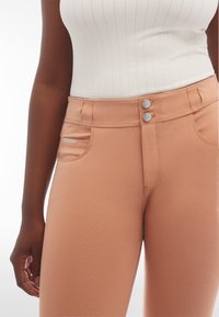 Pantalons couleur pêche en tissu extensible, dotés de deux boutons argentés à la taille et d'une petite poche latérale avec un logo brodé.
