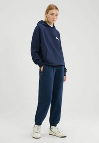 Granatowa bluza z kapturem z przednią kieszenią i logo, połączona z dopasowanymi zwężanymi dresami. Strój posiada elastyczne mankiety i miękką teksturę.