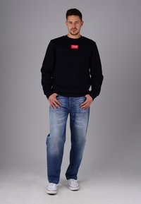 Black sweatshirt with a red logo patch, paired with light blue jeans and white sneakers. The fabric appears soft, and the fit is relaxed.