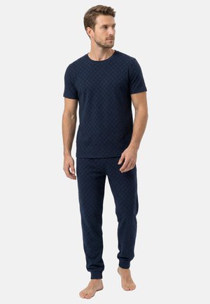 Mann steht barfuß und trägt ein passendes dunkelblaues kurzärmeliges Oberteil und Jogginghosen mit dezentem Muster vor einem einfarbigen Hintergrund.