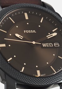 Schwarze Fossil-Uhr mit strukturiertem Gehäuse, rundem Ziffernblatt, goldfarbenen Zeigern und Datumsanzeige, die "MI 6" auf dunklem Hintergrund zeigt.