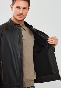 Chaqueta de cuero negro con detalles acolchados en los hombros, cierre de cremallera y forro interior suave, llevada sobre una camiseta polo beige.