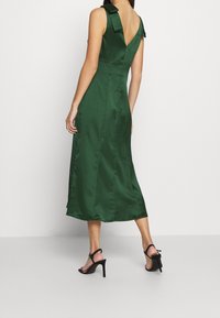 Nainen, joka on pukeutunut hihattomaan, syvän vihreään satiiniseen midi-mekkoon V-leikkaustakaa, yhdistettynä mustiin korkeakorkoisiin sandaaleihin, seisoo tasapohjalla.