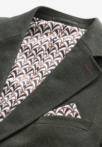 Blazer verde scuro con tessuto strutturato; interno caratterizzato da fodera a motivo bianca, marrone e nera. Il fazzoletto da taschino riprende il disegno della fodera.