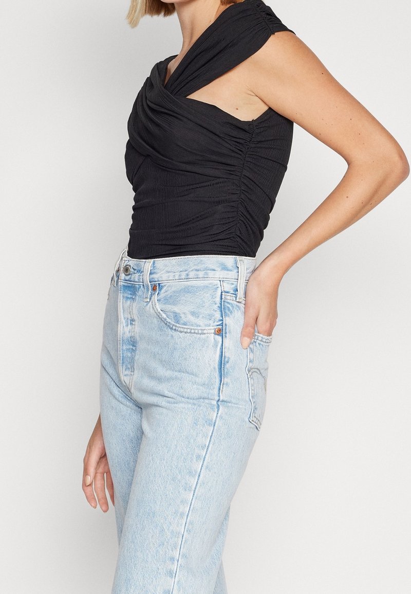 Černý nařasený top s draped výstřihem, zkombinovaný s světle modrými džínami s vysokým pasem. Látka má strukturovaný povrch.