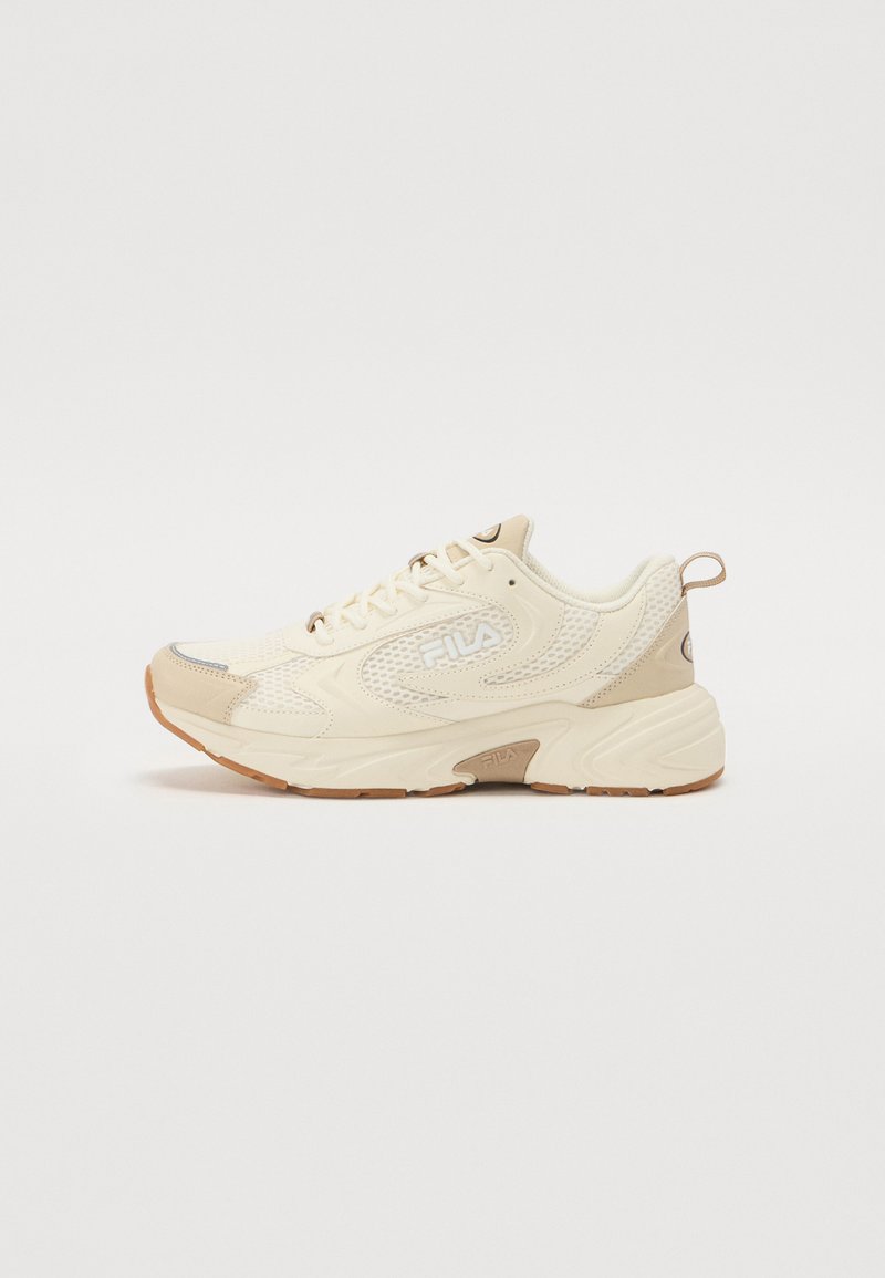 Zapatilla deportiva beige con parte superior de malla, suela acolchada y suela de goma. Presenta un logotipo en el lateral y una pestaña en el talón. Diseño de punta redondeada.