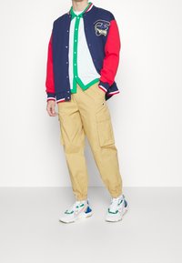 Marinblå och röd varsityjacka med broderad logotyp, vit skjorta med grön krage, beige cargobyxor och flerfärgade sneakers.