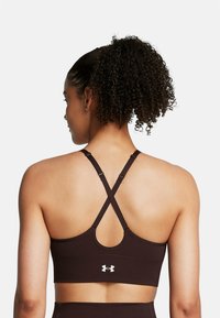 Bruine sportbeha met kruislingse bandjes, gestructureerd materiaal en een ronde uitsnijding aan de achterkant. Heeft een klein wit logo aan de basis.