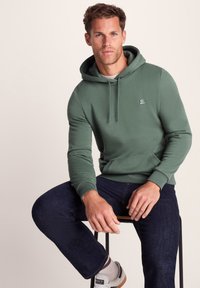 Man sitting on stool wearing green hoodie, dark jeans, white sneakers with striped socks, against plain beige background.