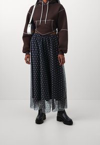 Brun cropped hoodie med hvid syning, parret med en gennemsigtig sort polkaprikker nederdel og sorte chunky støvler. Enkel sølvfarvet håndtaske.