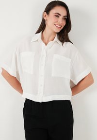White, short-sleeve blouse with a collar, featuring two chest pockets. Made of lightweight fabric with a boxy fit and button closure.