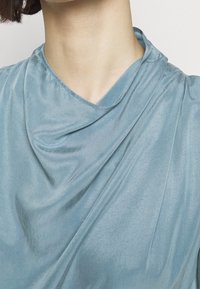 Nacke och skuldra på en person som bär en mjuk, ljusblå, sidenklänning med draperad halsringning och subtila veck mot en vit bakgrund.