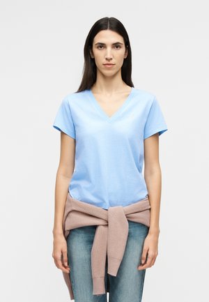 Hosszú, sötét hajú nő áll előre nézve, világoskék V-nyakú pólót, kék farmert és derekán megkötött rózsaszín pulóvert visel.