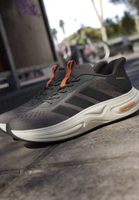 Paar graue und schwarze Laufschuhe mit orangefarbenen Zuglaschen, schwarzen Streifen und weißen gepolsterten Sohlen auf Außenpflaster.