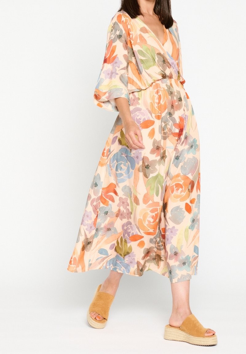 Femme en robe midi fluide à fleurs avec manches larges, portant des sandales compensées à enfiler couleur tan sur un fond blanc uni.