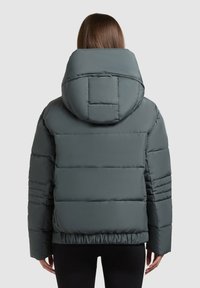 Person iført en grå gepolstret jakke med en stor hætte, vist fra ryggen mod en ensfarvet lys grå baggrund.