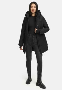 Parka nera con cappuccio, chiusura frontale a zip e bottoni automatici, con interni foderati e tasche laterali. Indossata sopra un top nero e jeans.