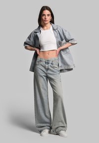 Denim jacket in light blue, oversized fit, paired with white cropped top and wide-leg light wash jeans. White sneakers complete the look.