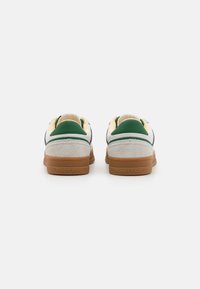 Zapatillas blancas con acentos verdes y talón de ante beige. Presentan una suela de goma y un logo de la marca visible en la parte trasera. Textura suave.