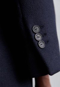 Gros plan sur une manche de manteau bleu marine avec trois boutons noirs gravés du nom de la marque "Sandro" cousus près du poignet.
