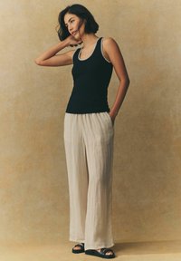 Sort tanktop med hvid kant, parret med beige brede bukser. Outfittet har en afslappet pasform og blødt, struktureret stof. Sorte sandaler fuldender looket.