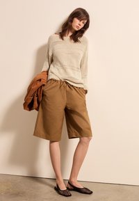 Beige Strickpullover mit oversize Schnitt, kombiniert mit braunen knielangen Shorts und schwarzen Netzslippers. Der Hintergrund ist schlicht.