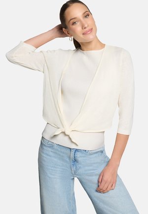 Femme aux cheveux châtain clair portant un cardigan blanc noué sur le devant, un haut blanc côtelé et un jean bleu clair, posant avec une main derrière la tête.