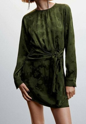Long-sleeve dark green floral dress with a tied waist sash, worn by a person with short hair and dangling chain earrings.