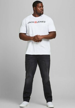 White short-sleeve t-shirt with "JACK & JONES" logo in black and red; paired with dark jeans and white sneakers. Model standing with arms crossed.